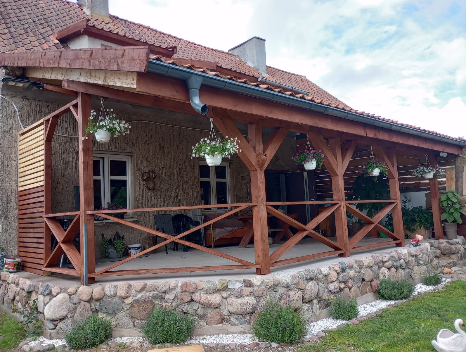 Zadaszony taras z drewnianą konstrukcją, ozdobiony wiszącymi kwiatami, z widocznym fragmentem dachu z dachówką ceramiczną i kamienną podmurówką.