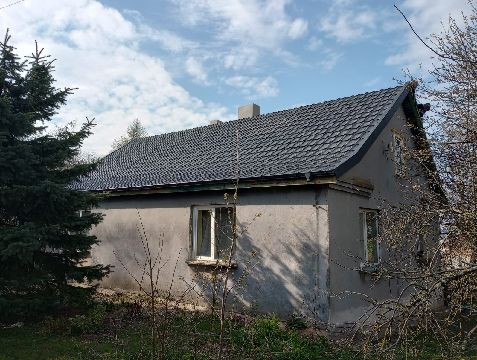 Dom jednorodzinny z nowym, grafitowym dachem z dachówki bitumicznej, widoczny z boku, z fragmentami drzew iglastych i liściastych w kadrze.