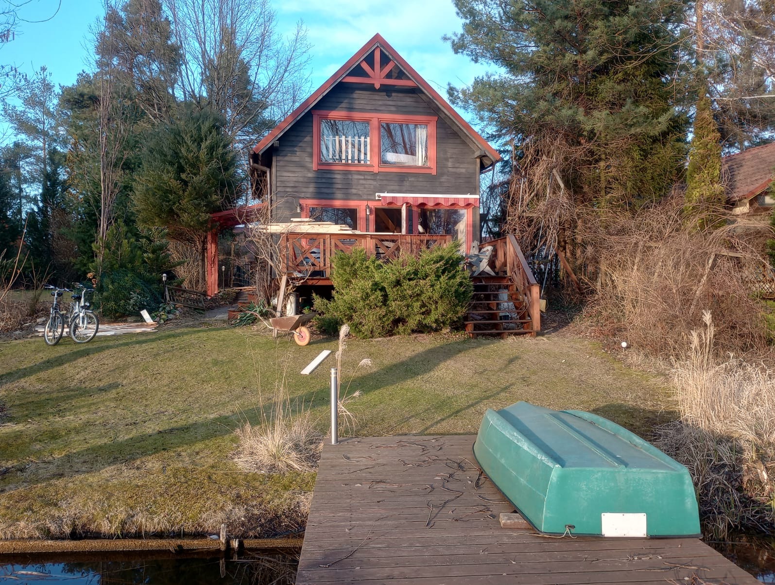 Drewniany dom z czerwonymi akcentami, widok z pomostu z odwróconą łódką, na trawniku rowery i taczka.
