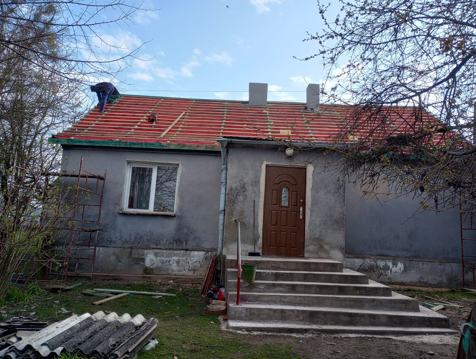 Pracownik na dachu szarego domu jednorodzinnego, montaż łat pod czerwone dachówki, widoczne rusztowanie i stos starych płyt eternitowych na trawniku.