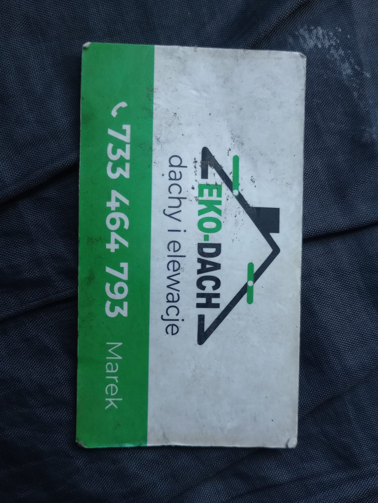 Wizytówka firmy EKO-DACH oferującej usługi w zakresie dachów i elewacji, z numerem telefonu 733 464 793 i imieniem Marek, leżąca na ciemnym materiale.