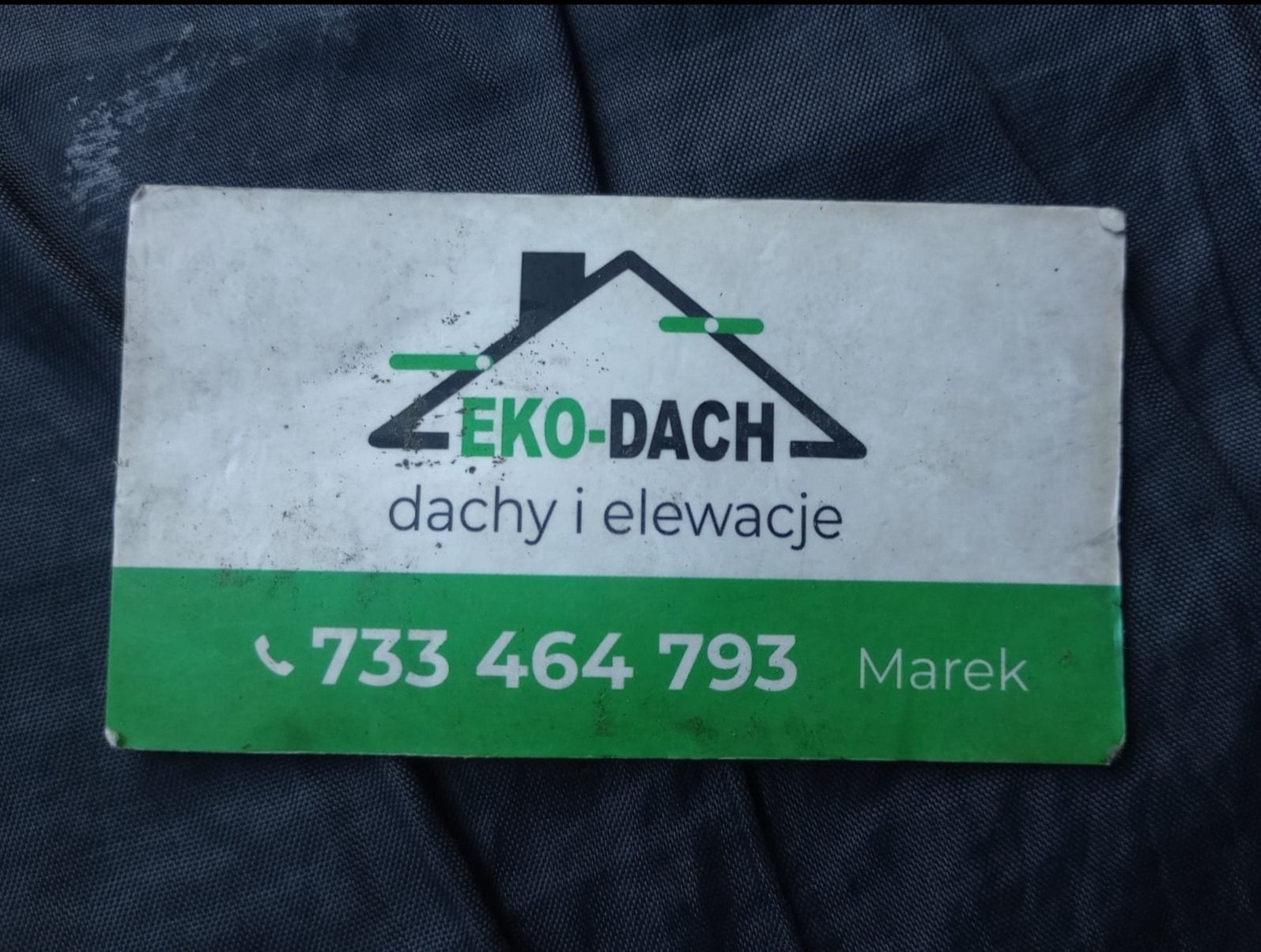 Wizytówka firmy EKO-DACH oferującej usługi związane z dachami i elewacjami, z widocznym numerem telefonu 733 464 793 i imieniem Marek, leżąca na ciemnym materiale.
