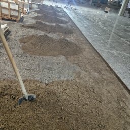Spec bud Paweł Żak - Przygotowanie podłoża z piasku pod płytki w dużej hali, widoczne palety z płytkami i ekipa montażowa w tle.