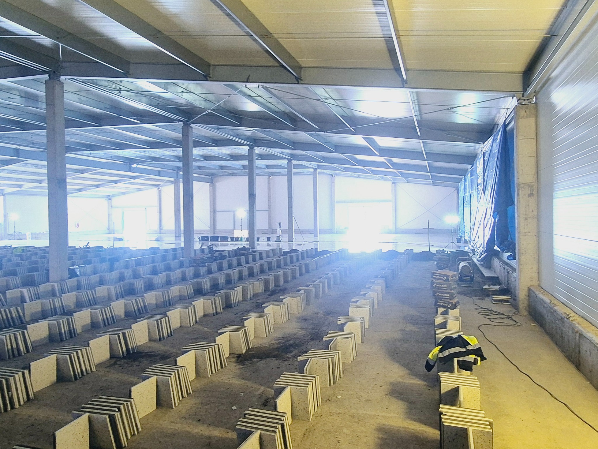 Stosy płytek podłogowych ułożone w rzędach w hali przemysłowej przed ułożeniem, widoczne narzędzia i odzież robocza w tle.