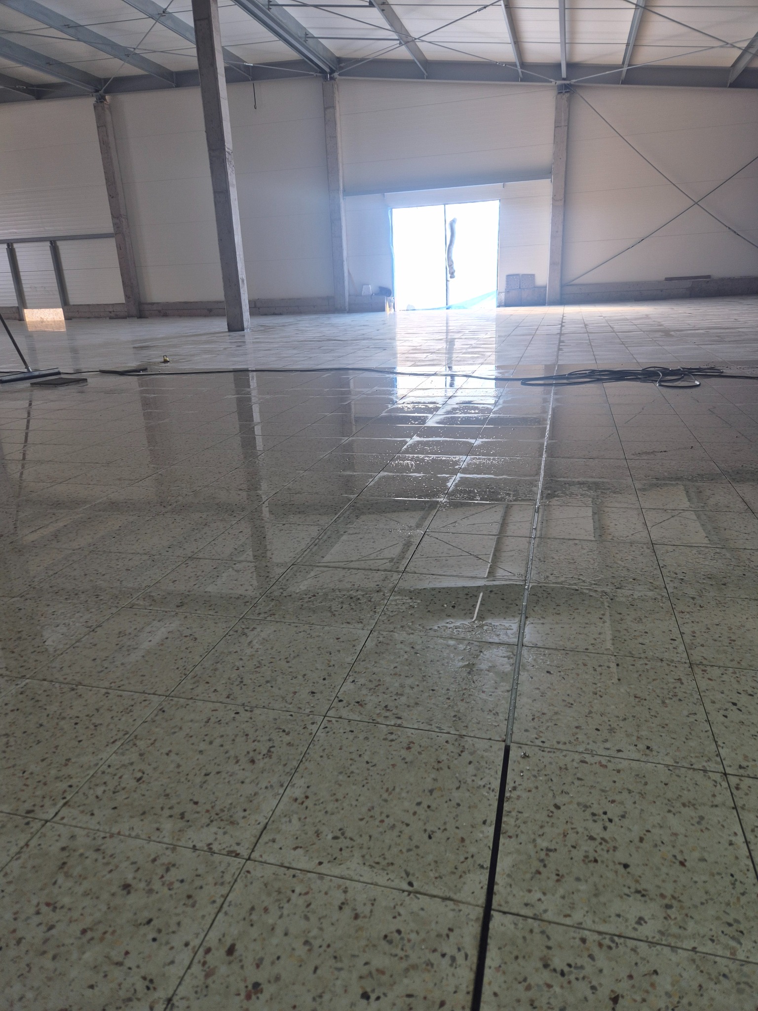 Świeżo położona, mokra podłoga z płytek w dużej hali, z widocznymi fugami i odbiciami światła, gotowa do dalszych prac wykończeniowych.