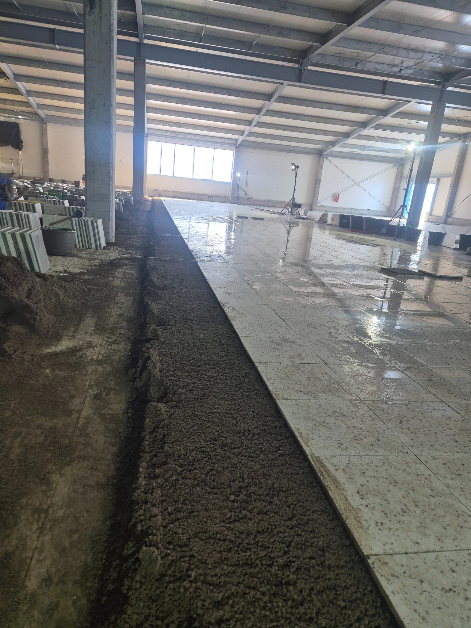 Układanie płytek podłogowych w hali przemysłowej, widoczny fragment świeżo ułożonych płytek i przygotowane podłoże z zaprawą, oświetlenie budowlane w tle.