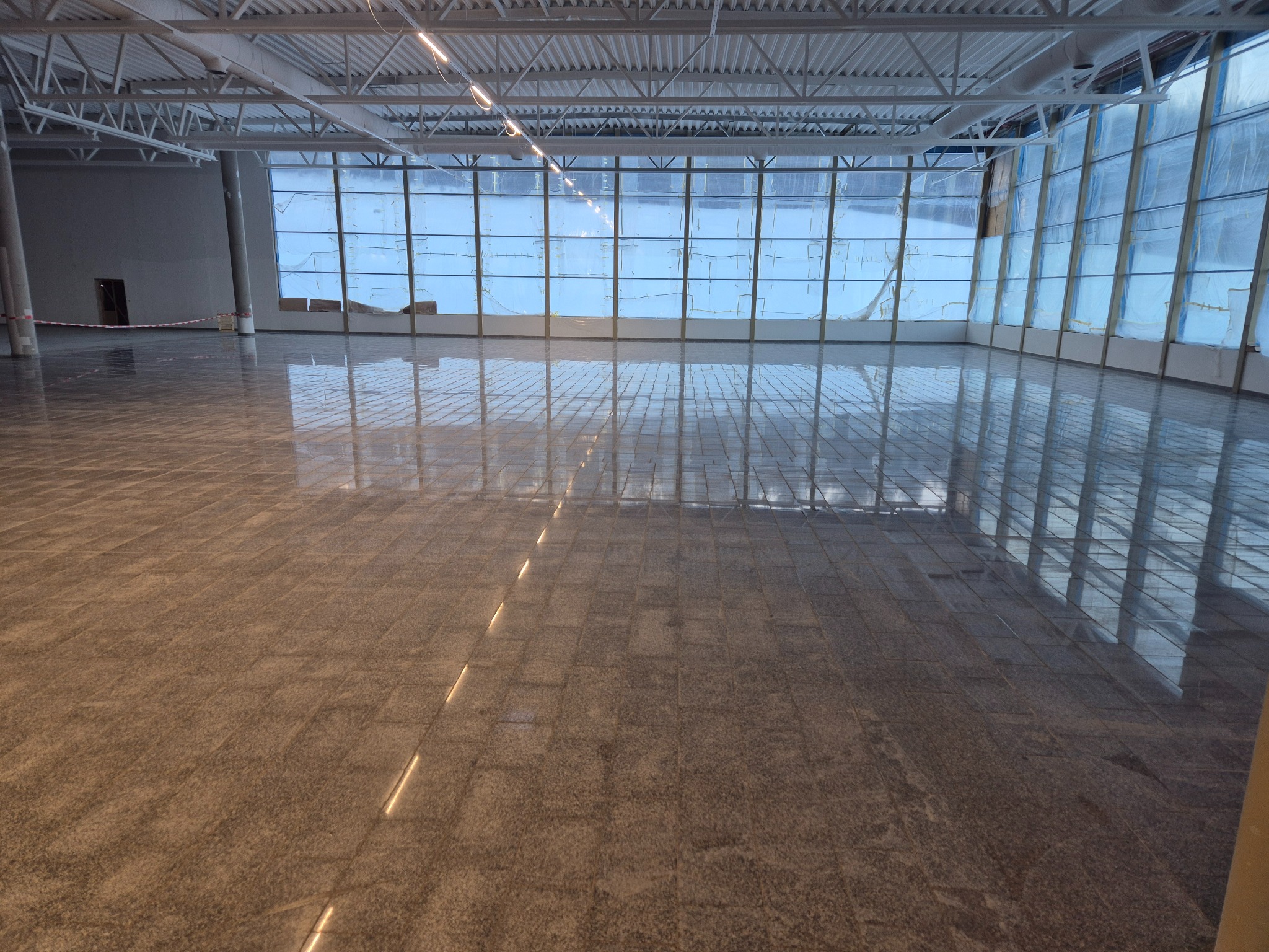 Błyszcząca posadzka z szarej terakoty w nowym, przestronnym pomieszczeniu z dużymi oknami i stalową konstrukcją dachu. Podłoga odbija światło, tworząc efekt lustrzany.