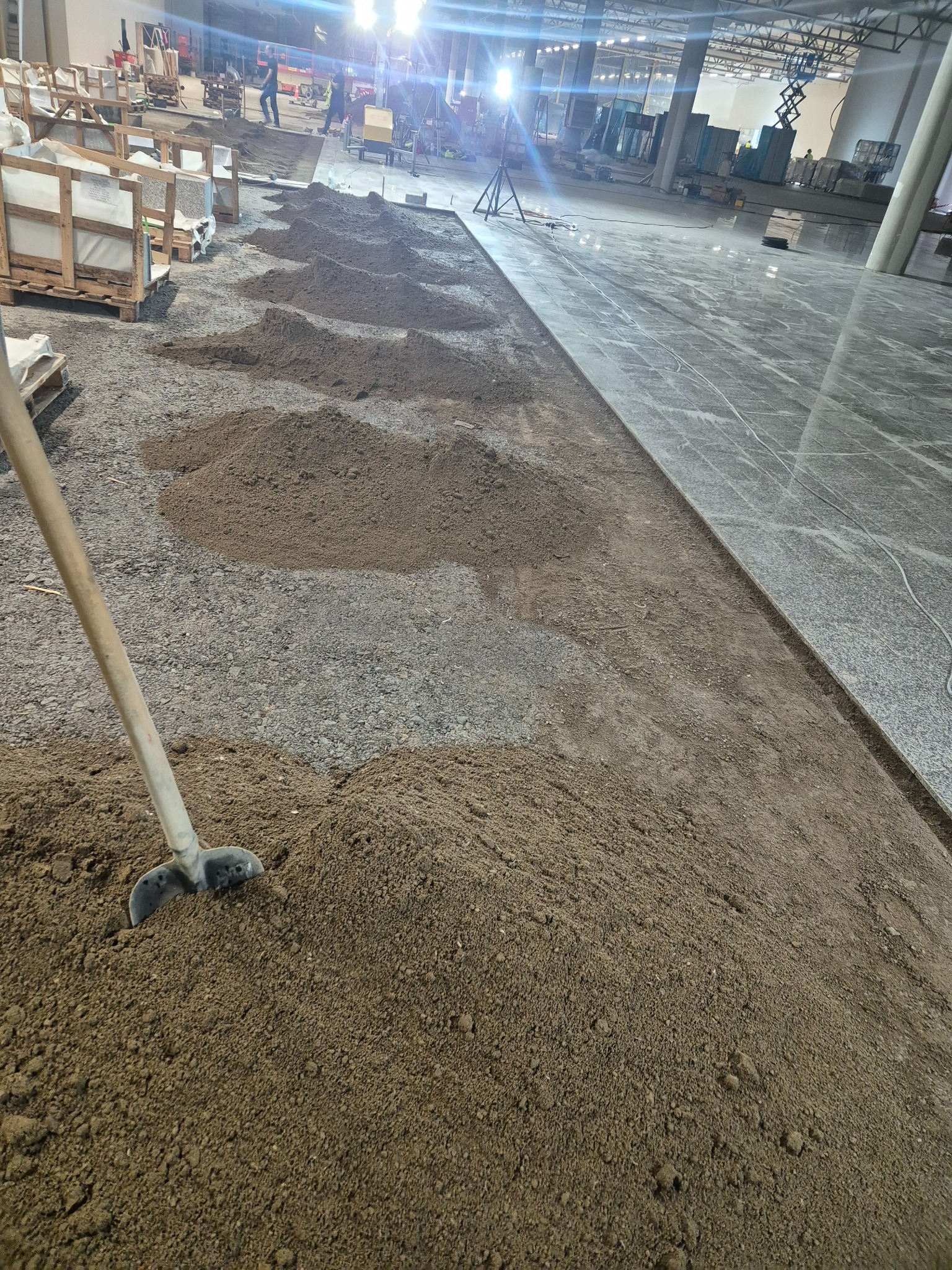 Przygotowanie podłoża z piasku pod płytki w dużej hali, widoczne palety z płytkami i ekipa montażowa w tle.
