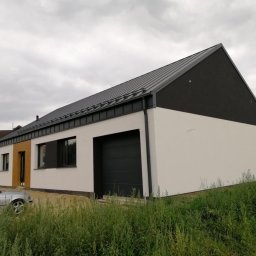 DOMINBUD IK SPÓŁKA Z OGRANICZONĄ ODPOWIEDZIALNOŚCIĄ - Nowoczesny dom jednorodzinny z garażem, wykończony w bieli i czerni, z metalowym dachem. Widok z poziomu gruntu, zarośnięty teren przed budynkiem.