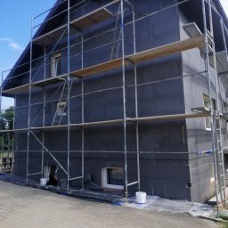 DOMINBUD IK SPÓŁKA Z OGRANICZONĄ ODPOWIEDZIALNOŚCIĄ - Dom w trakcie ocieplania, widoczne rusztowania i płyty izolacyjne na elewacji. Prace w toku, z detalami jak wiaderka i zabezpieczenia na ziemi.