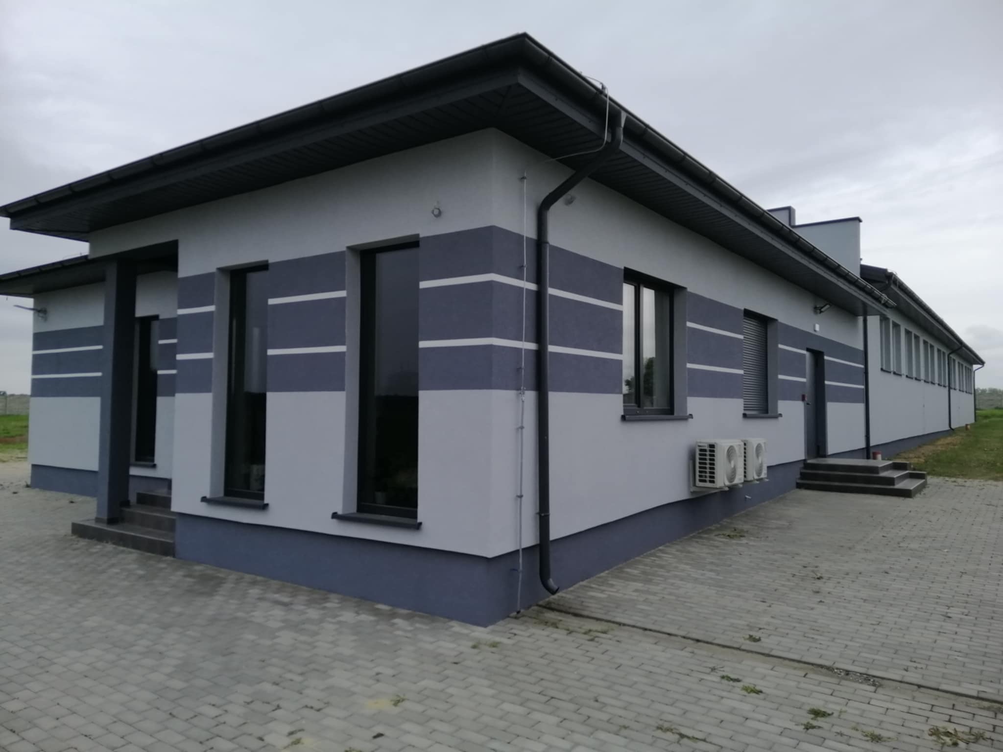 Nowoczesny budynek z szarą elewacją, ciemnymi akcentami i poziomymi pasami. Widoczne okna, klimatyzatory i kostka brukowa. Pochmurne niebo w tle.