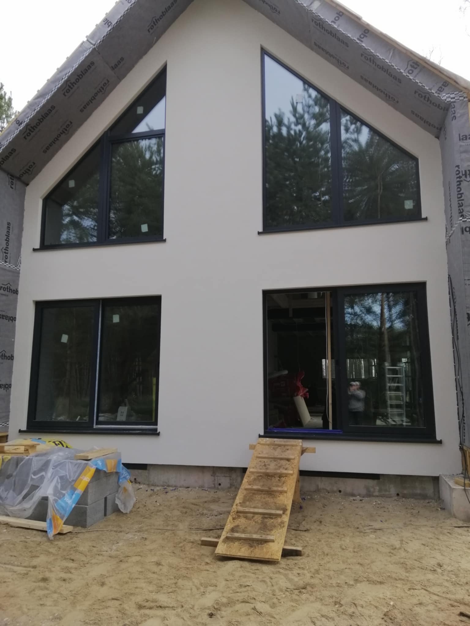 Nowoczesny dom z jasną elewacją i dużymi oknami o nietypowym kształcie. Widoczne materiały budowlane i rusztowanie, sugerujące prace wykończeniowe.