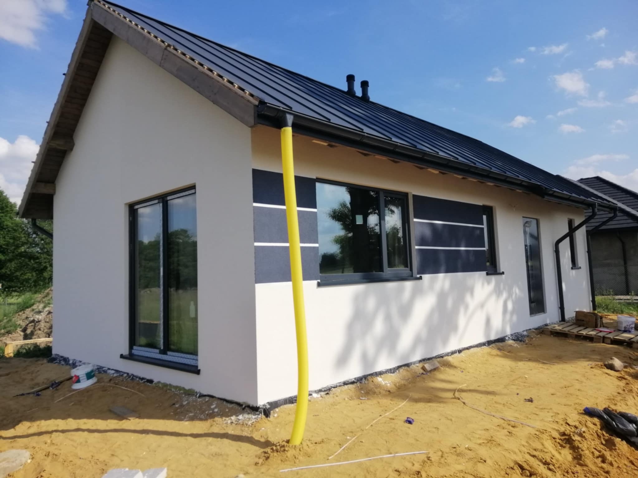 Nowoczesny, parterowy dom z białą elewacją i grafitowymi akcentami, czarnym dachem. Żółta rura spustowa na pierwszym planie. Widoczne okna i drzwi.