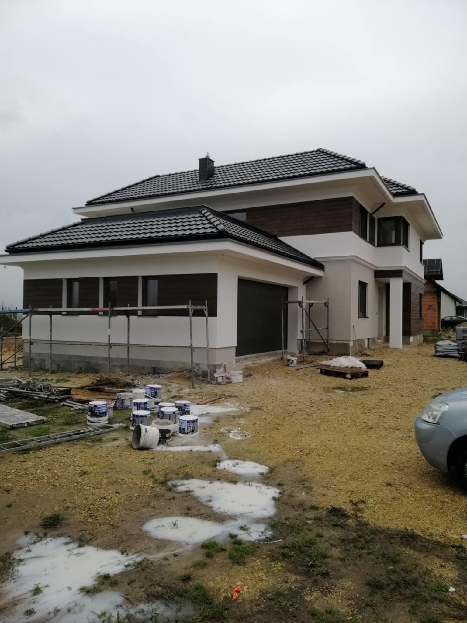 Dom jednorodzinny w trakcie malowania elewacji, widoczne rusztowania i puszki z farbą. Częściowo wykończona fasada z elementami drewnianymi. Pochmurny dzień.