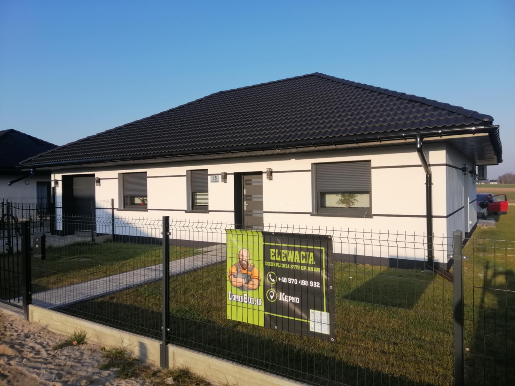 Dom jednorodzinny z jasną elewacją i ciemnym dachem, ogrodzony czarnym płotem. Na płocie baner reklamowy firmy budowlanej z hasłem 'Elewacja'.