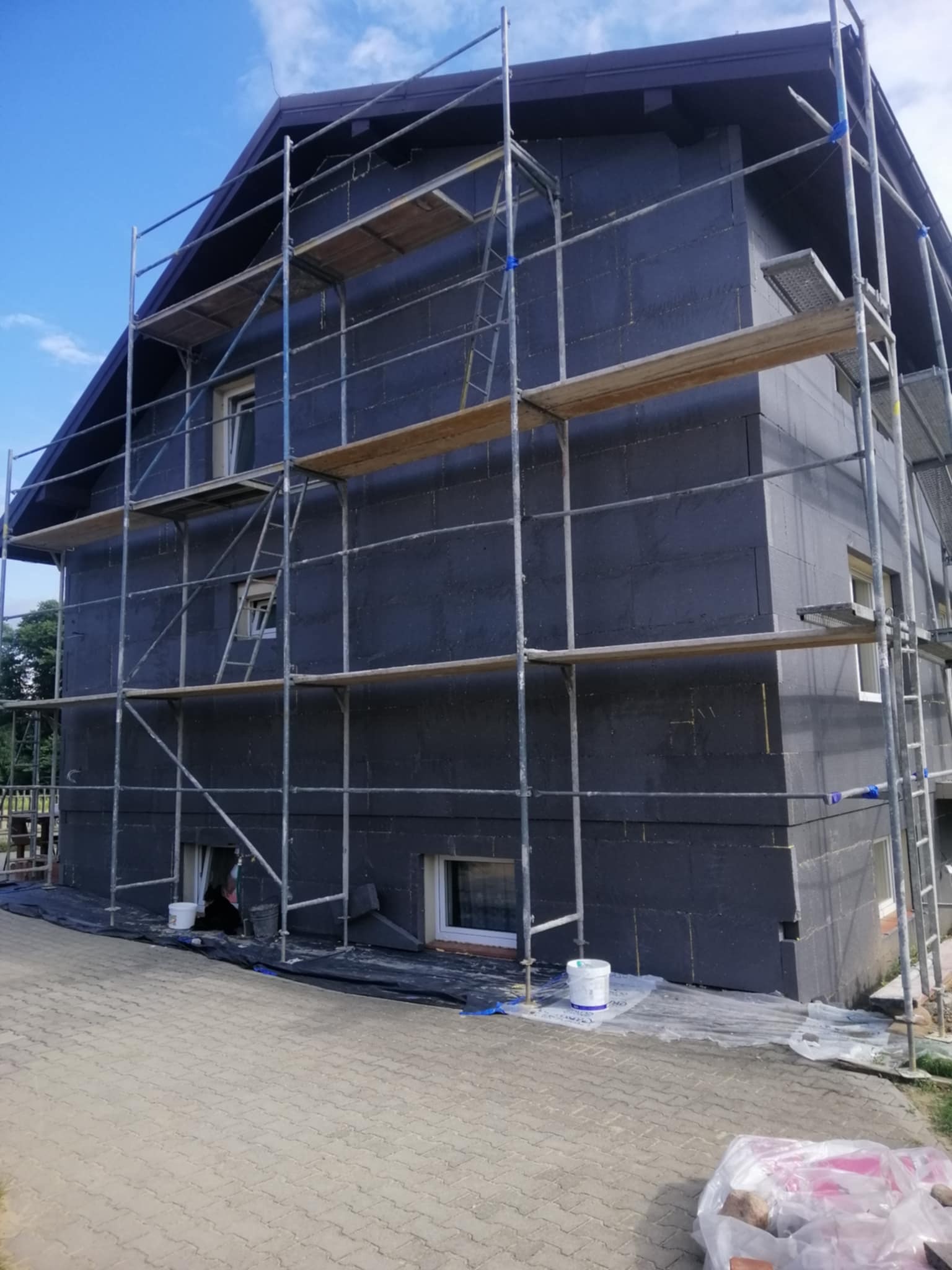 Dom w trakcie ocieplania, widoczne rusztowania i płyty izolacyjne na elewacji. Prace w toku, z detalami jak wiaderka i zabezpieczenia na ziemi.