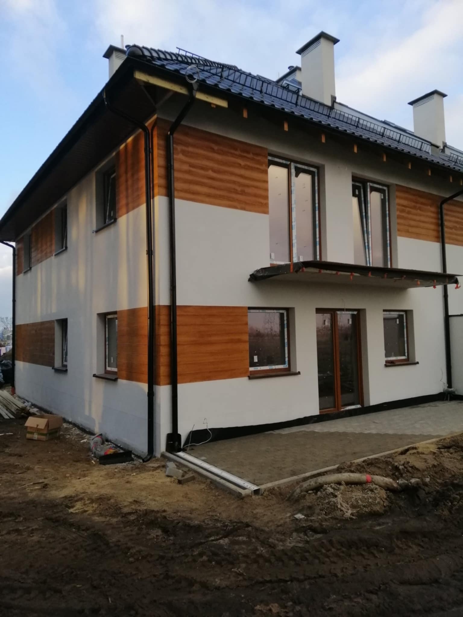 Dwukondygnacyjny dom z elewacją w poziome pasy: biały tynk i drewnopodobne panele. Widoczne rynny, okna z brązowymi ramami i częściowo wykończony teren wokół.