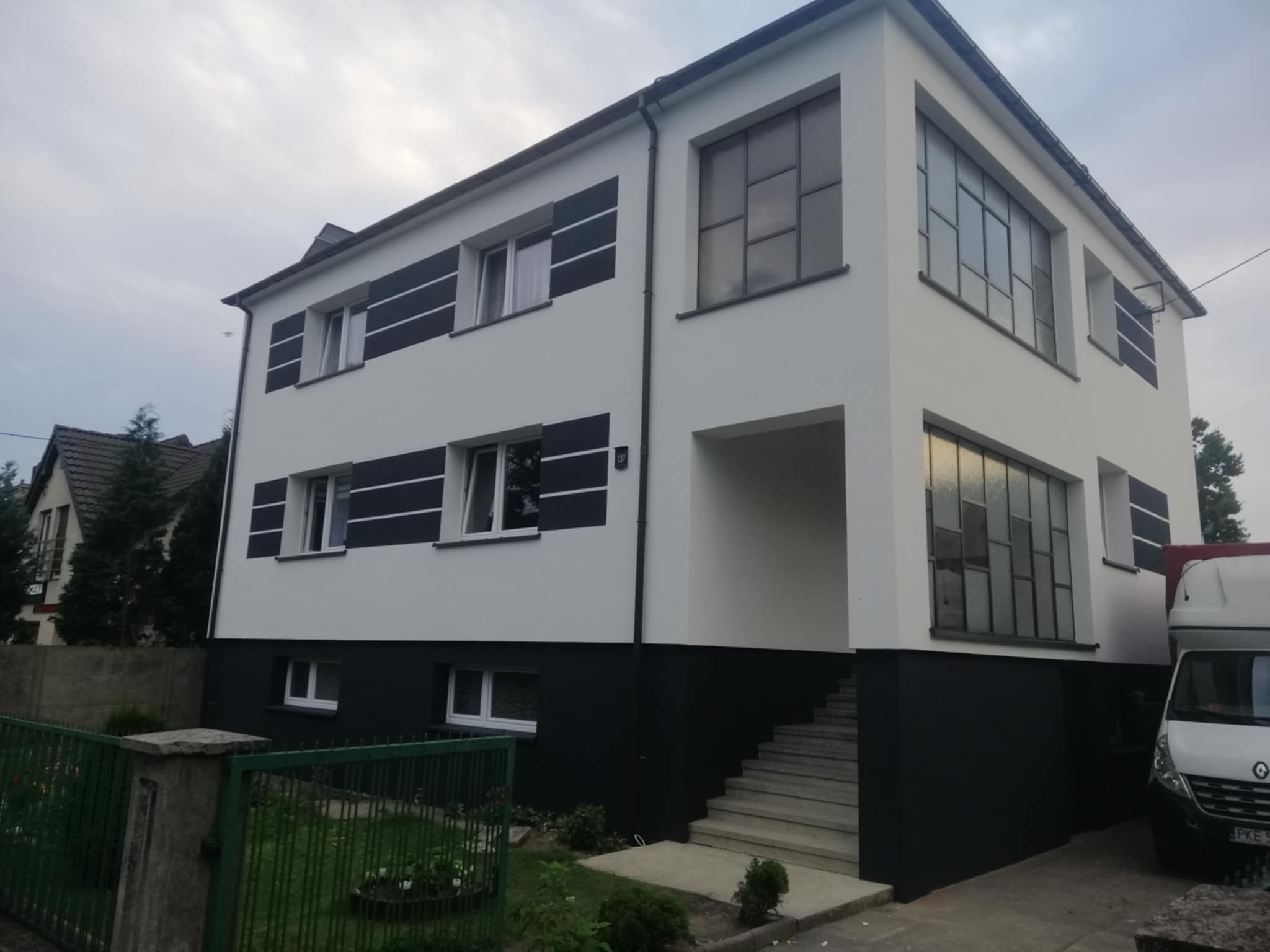 Dwukondygnacyjny dom po renowacji elewacji, z czarno-białą kolorystyką i dużymi oknami, widziany z perspektywy ogrodu z zielonym ogrodzeniem.