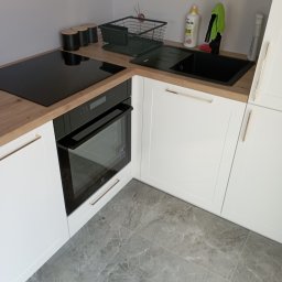 Tomasz Kuna Strefa Mebli - Fragment nowoczesnej kuchni z białymi szafkami z frezowanymi frontami i złotymi uchwytami, drewnianym blatem, czarną płytą indukcyjną i piekarnikiem oraz szarymi płytkami podłogowymi imitującymi...