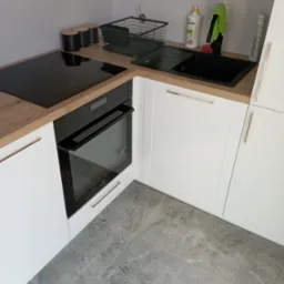 Fragment nowoczesnej kuchni z białymi szafkami z frezowanymi frontami i złotymi uchwytami, drewnianym blatem, czarną płytą indukcyjną i piekarnikiem oraz szarymi płytkami podłogowymi imitującymi...