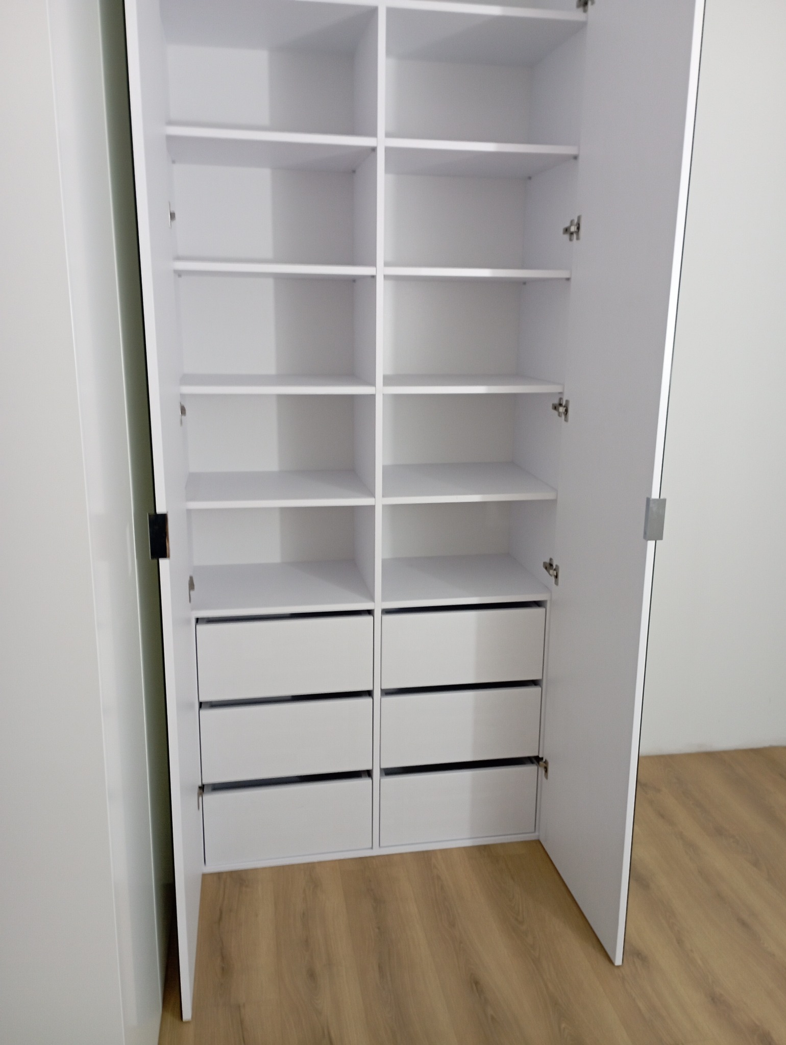 Biała szafa wnękowa z otwartymi drzwiami, widoczne półki i szuflady, minimalistyczny design, jasne wnętrze, drewniana podłoga.