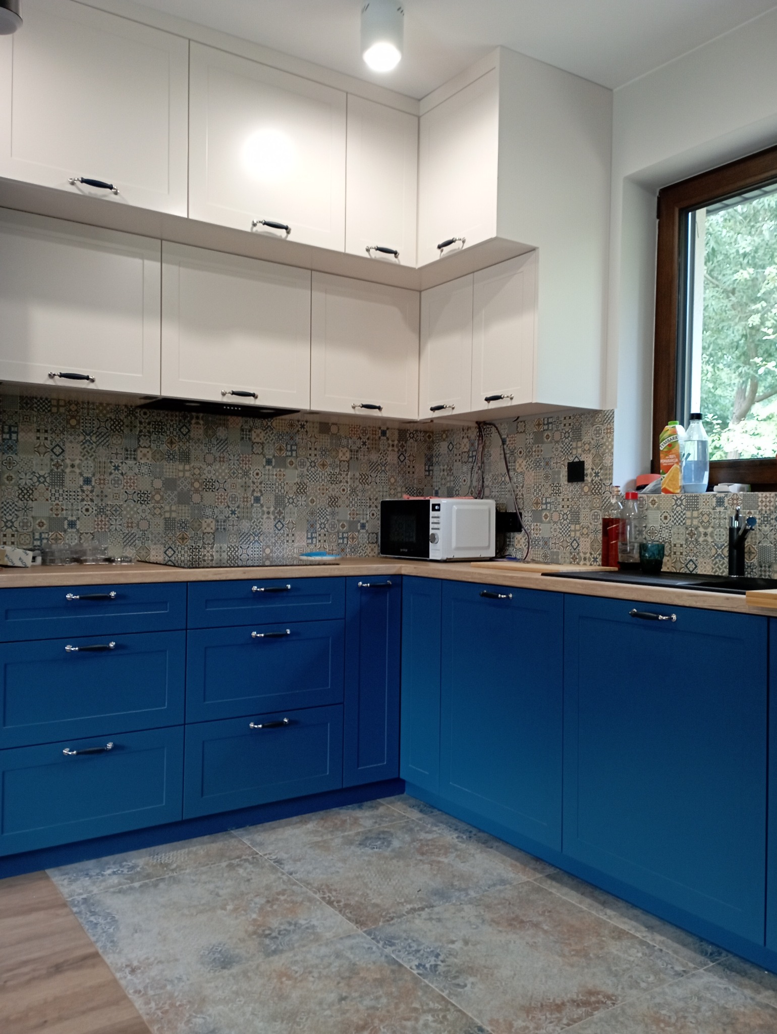 Niebiesko-biała kuchnia z patchworkowymi płytkami nad blatem i drewnianym oknem z widokiem na zieleń.