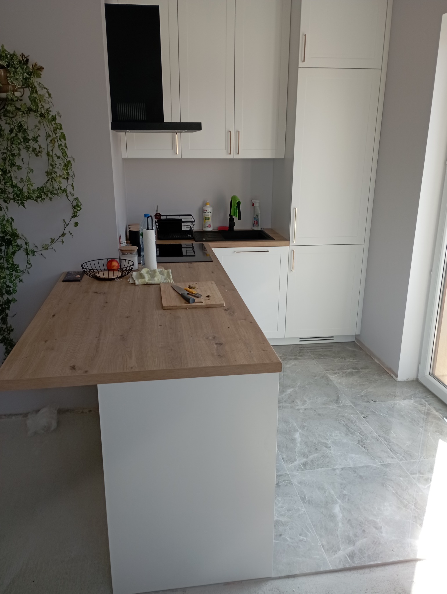 Nowoczesna, biała kuchnia z drewnianym blatem roboczym, na którym leżą nóż i deska do krojenia. Na blacie widoczny również kosz z jabłkiem i drobne akcesoria kuchenne. W tle czarny okap i marmurowe...