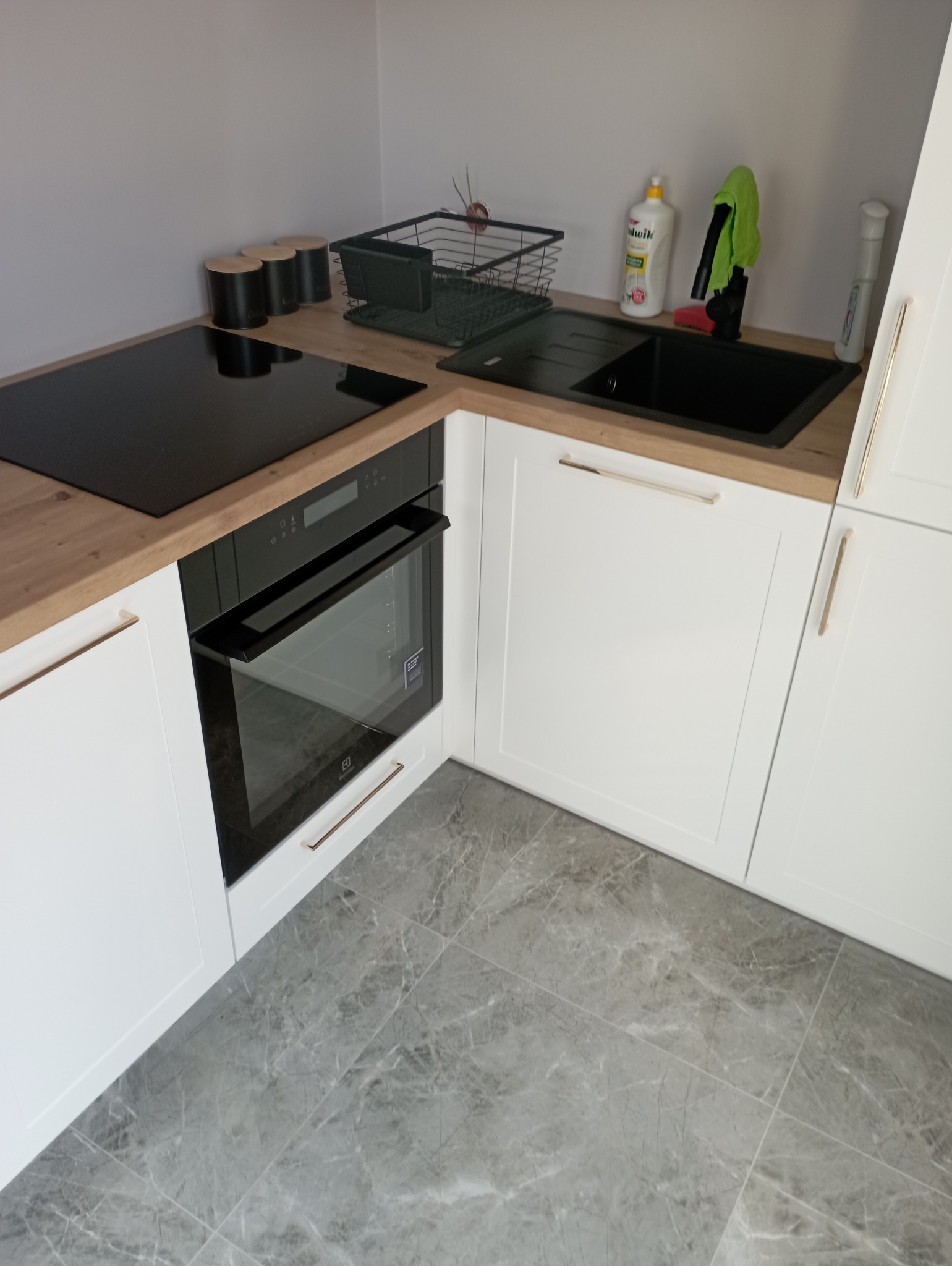 Fragment nowoczesnej kuchni z białymi szafkami z frezowanymi frontami i złotymi uchwytami, drewnianym blatem, czarną płytą indukcyjną i piekarnikiem oraz szarymi płytkami podłogowymi imitującymi...