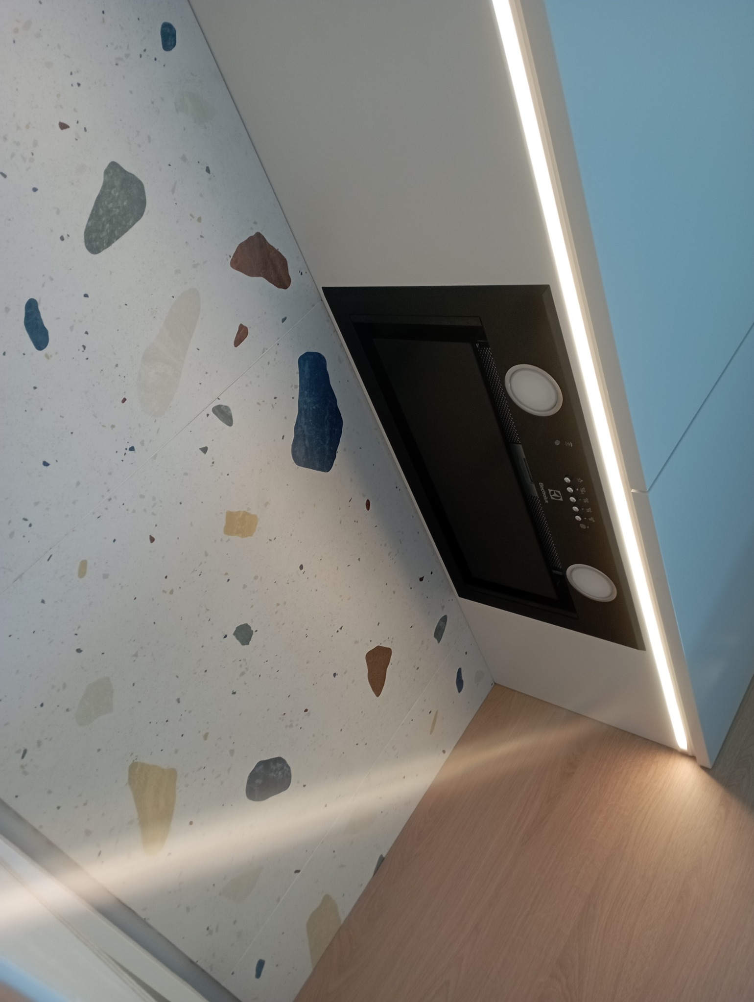 Fragment kuchni na wymiar: okap Electrolux podświetlony listwą LED, ściana wykończona białą płytką z kolorowymi plamkami, drewnopodobny blat.