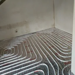 Białe rury ogrzewania podłogowego marki Roth ułożone na folii z czerwoną kratką w surowym wnętrzu z nieotynkowanym sufitem, widok z góry pod kątem.