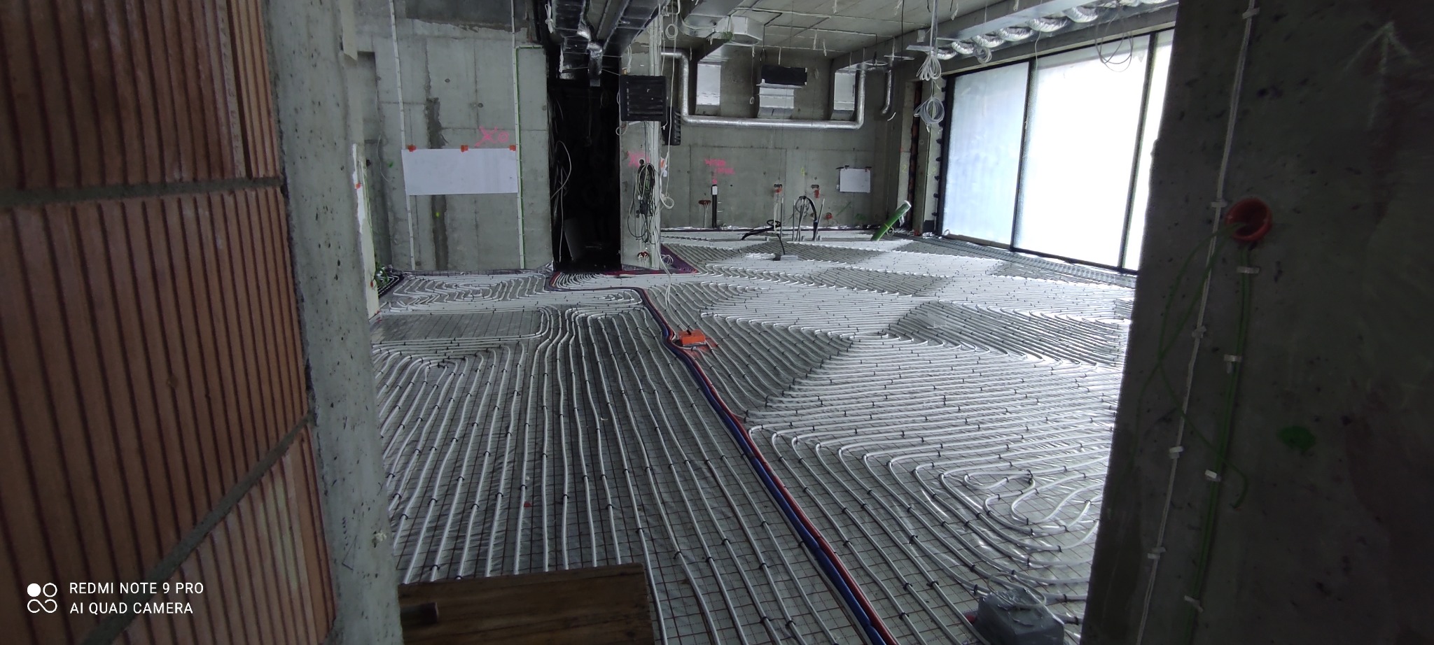 Rozłożone białe rurki ogrzewania podłogowego na siatce w surowym wnętrzu budynku w trakcie budowy, widoczne betonowe ściany i instalacje pod sufitem.