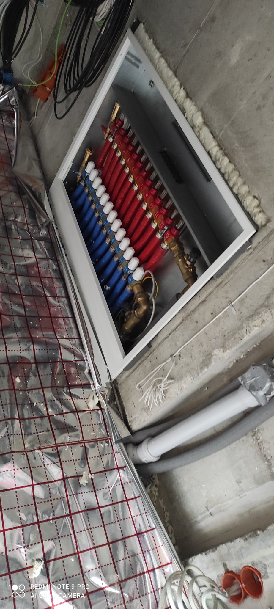 Rozdzielacz ogrzewania podłogowego w szafce, widoczne rury czerwone i niebieskie z numeracją, izolacja podłogowa z folii aluminiowej w tle.