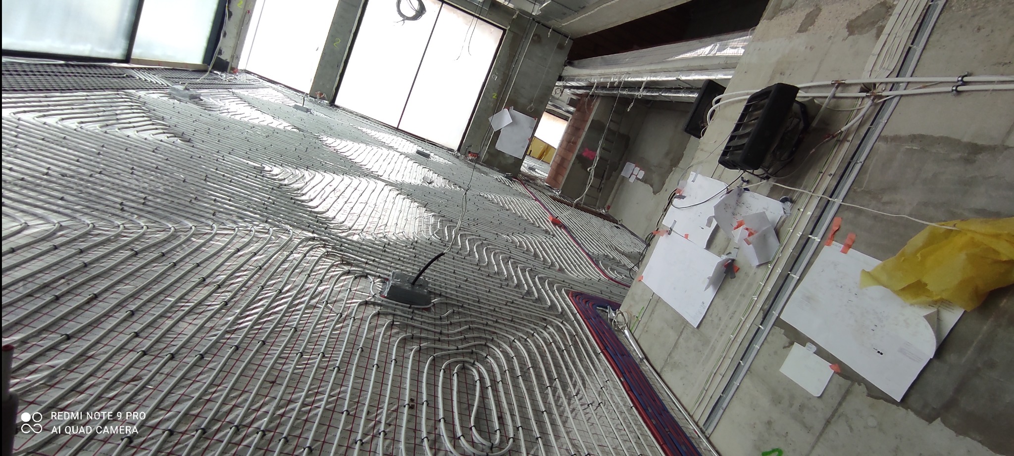 Układ rur ogrzewania podłogowego w surowym wnętrzu, widoczne białe rury ułożone na folii, czerwona siatka montażowa, okno w tle i plany na ścianie.