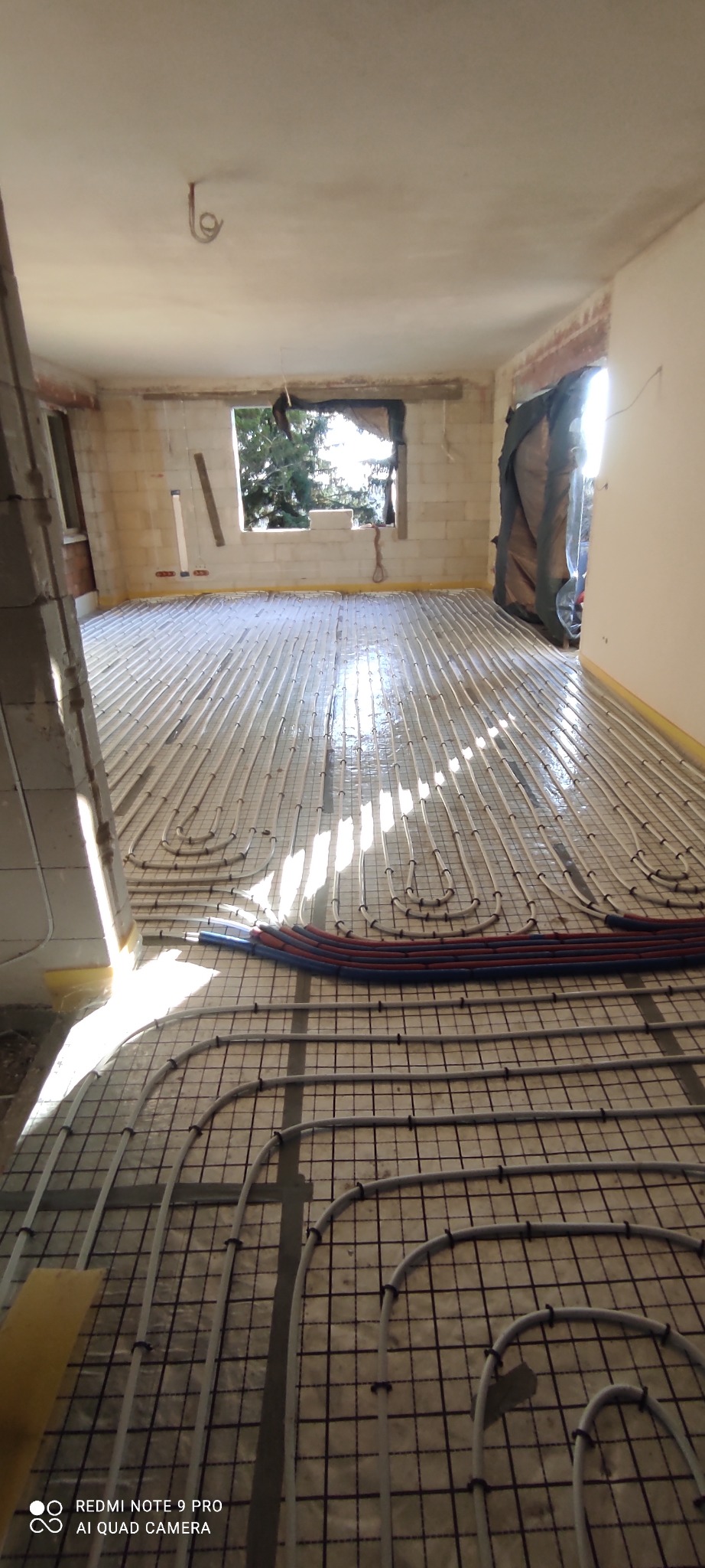 Wnętrze w trakcie montażu ogrzewania podłogowego: rury grzewcze ułożone na siatce, widoczne okno i surowe ściany.