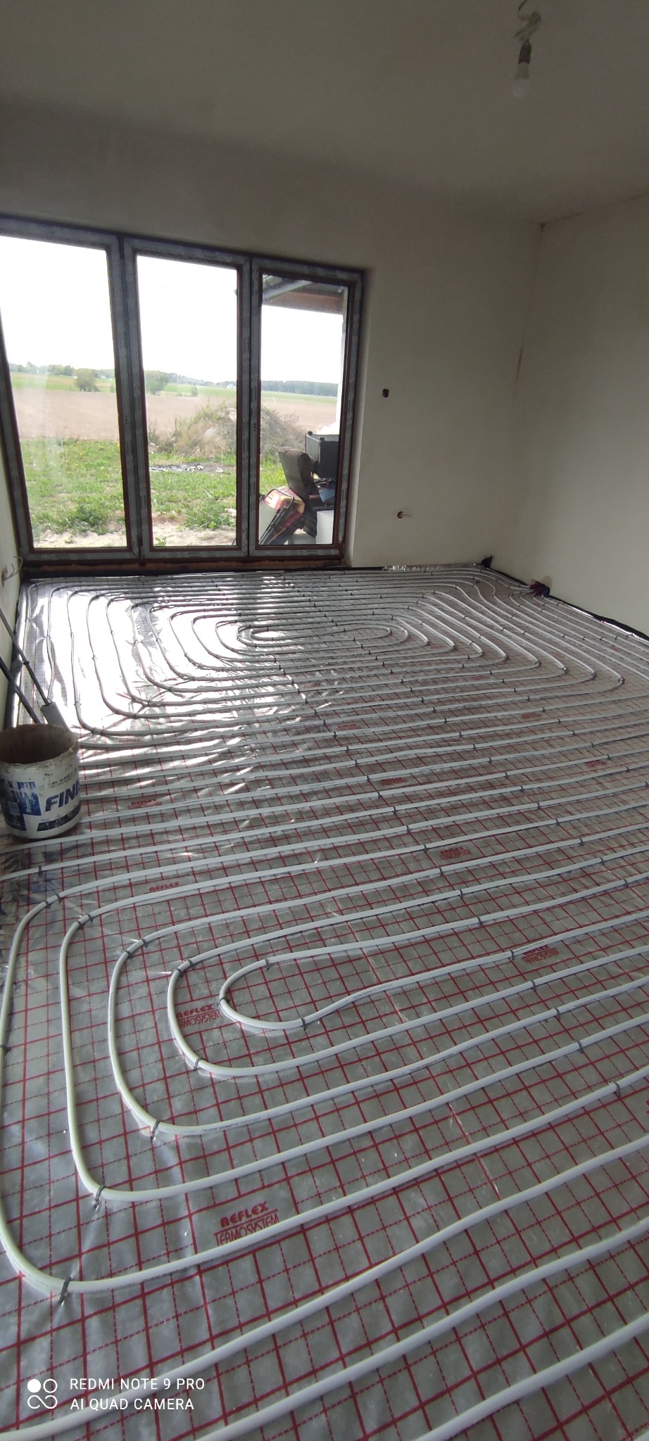 Białe rury ogrzewania podłogowego ułożone spiralnie na podkładzie z czerwoną siatką w surowym pomieszczeniu z oknami wychodzącymi na pole.