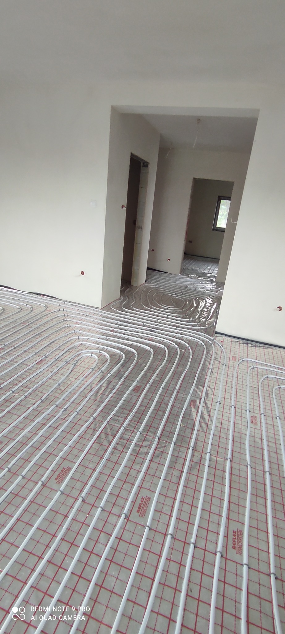 Białe rurki ogrzewania podłogowego ułożone na czerwonej siatce w surowym wnętrzu z widocznymi otworami drzwiowymi.