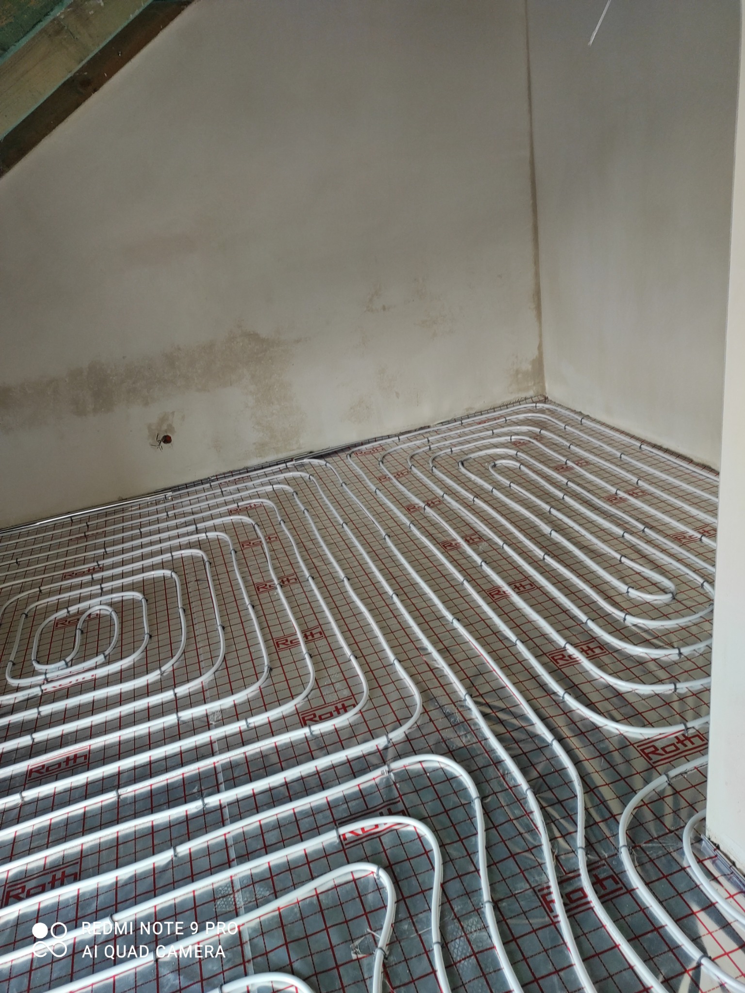 Białe rury ogrzewania podłogowego marki Roth ułożone na folii z czerwoną kratką w surowym wnętrzu z nieotynkowanym sufitem, widok z góry pod kątem.