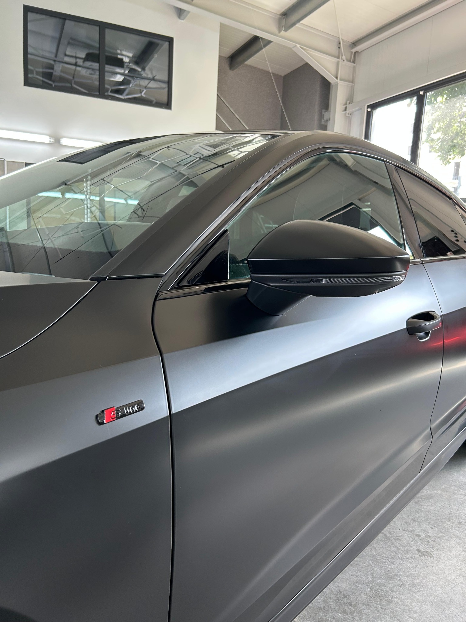 Matowy, grafitowy Audi A-line po zmianie koloru folią. Widoczne detale: lusterko, klamka, emblemat. Auto w jasnym garażu.