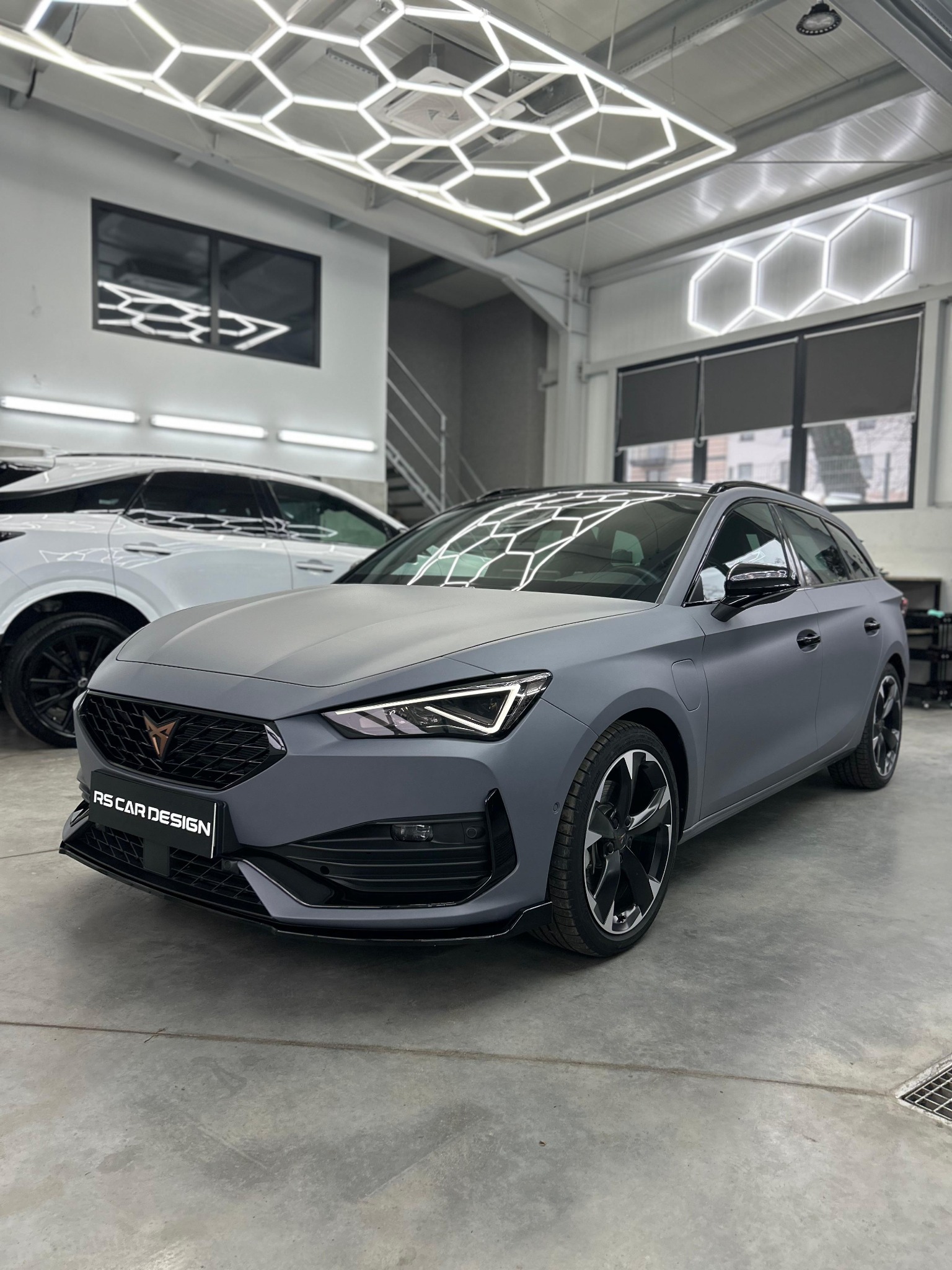 Szary samochód Cupra Leon po oklejeniu folią matową w studiu RS Car Design, z widocznym logo firmy. W tle biały SUV i heksagonalne oświetlenie sufitowe.