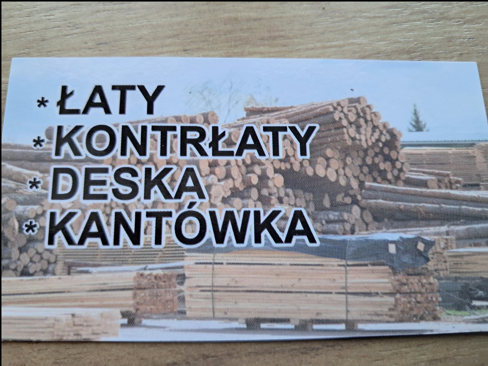 Wizytówka z listą produktów: łaty, kontrłaty, deska, kantówka, na tle stosów drewna.