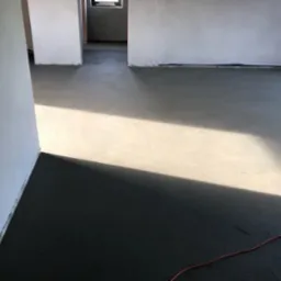 Świeżo wylana, gładka posadzka w surowym wnętrzu z widocznym promieniem słonecznym i fragmentem czerwonego kabla.