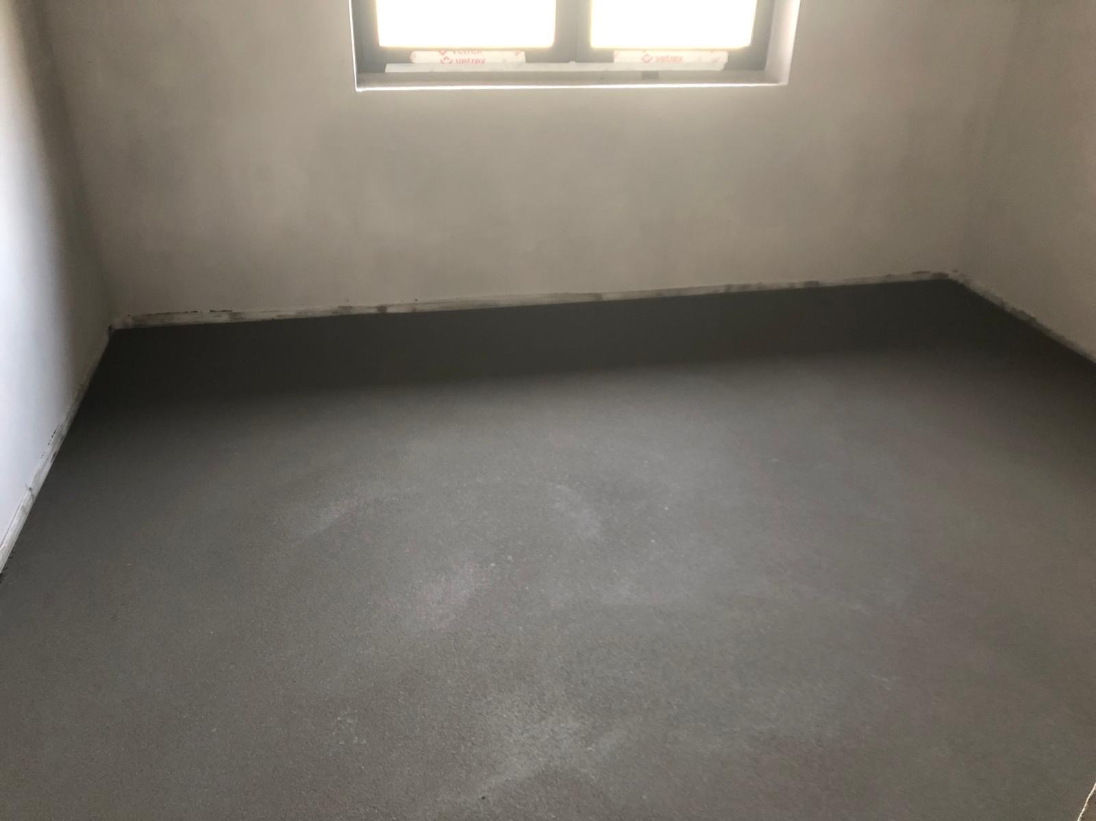 Świeżo wylana, szara wylewka podłogowa w pomieszczeniu z oknem, widoczne ślady po poziomowaniu.