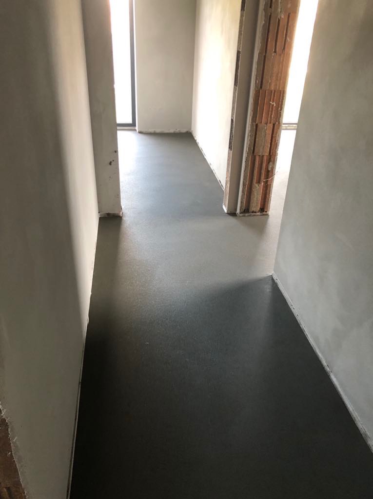 Świeżo wylana, gładka, ciemnoszara posadzka w korytarzu z widocznymi surowymi ścianami i fragmentem niezabudowanej ościeżnicy z cegły dziurawki.