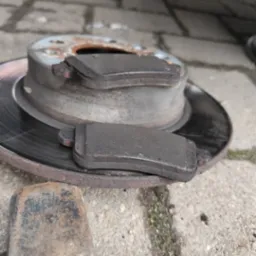 Zużyte klocki hamulcowe leżące na tarczy hamulcowej obok metalowego młotka na betonowej kostce brukowej.