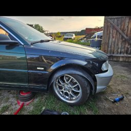 Jakub Pakulski - Fragment samochodu z widocznym podnośnikiem pneumatycznym, narzędziami i uszkodzonym błotnikiem, sugerujący prace naprawcze w warunkach warsztatowych.