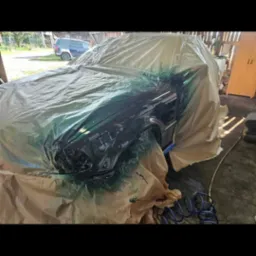 Samochód częściowo zakryty folią ochronną przed lakierowaniem, widoczne ślady farby na folii i elementach auta, niebieski wąż pneumatyczny leży obok.