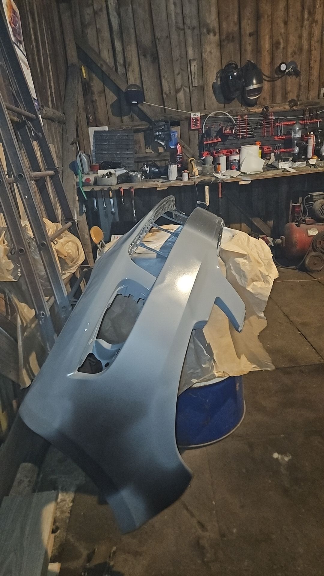 Szary, świeżo polakierowany zderzak samochodowy, ustawiony na niebieskim beczce w warsztacie z drabina i narzędziami w tle.