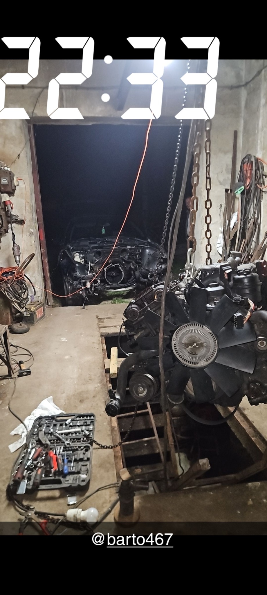 Demontaż silnika samochodowego w warsztacie: silnik zawieszony na łańcuchu nad wykopem, skrzynka z narzędziami obok, w tle widoczny zdekompletowany samochód.