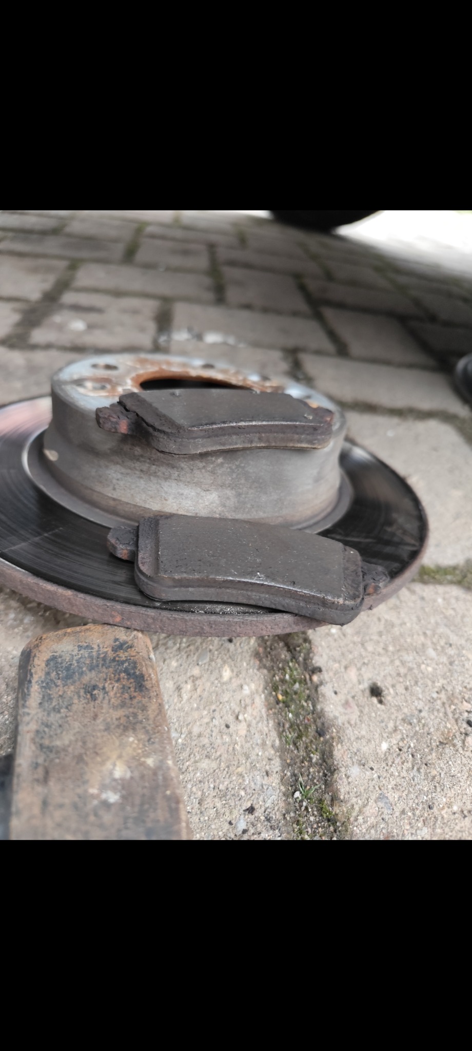 Zużyte klocki hamulcowe leżące na tarczy hamulcowej obok metalowego młotka na betonowej kostce brukowej.