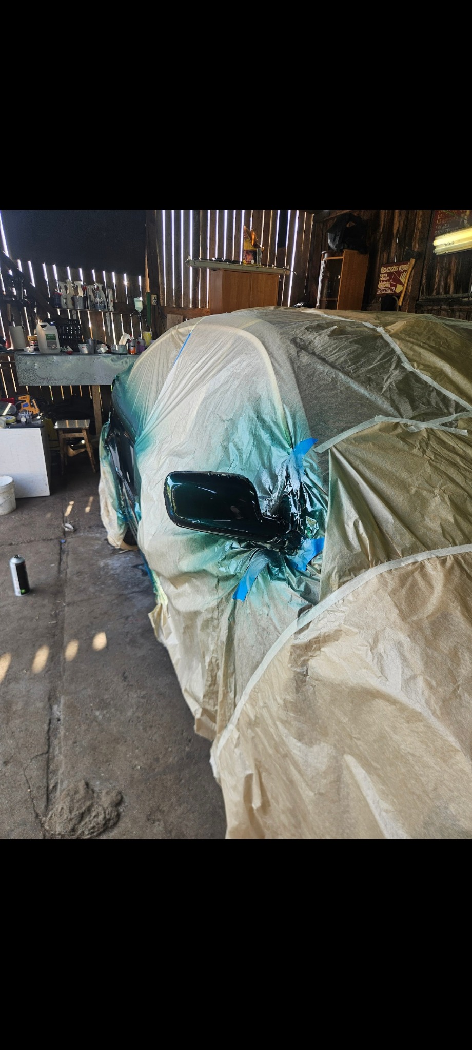 Samochód częściowo zakryty folią ochronną podczas lakierowania w warsztacie, z widocznym lusterkiem i świeżą warstwą lakieru, obok puszka z farbą w sprayu.